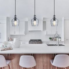 three lights hanging over a kitchen island with white cabinets and stools next to it