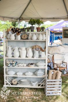 a white shelf filled with lots of plates and cups on top of wooden crates under a tent