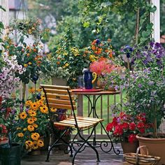 a table and chair on a deck with flowers in the back ground, next to potted plants