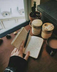 a person writing on an open book next to some coffee cups and jars with plants in them