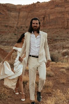 a man and woman walking through the desert holding hands, dressed in all white clothes