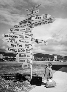 black and white photograph of a person standing next to a street sign with many directions on it