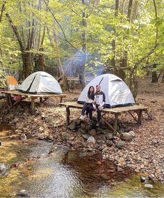 two people sitting on a picnic table next to some tents in the woods near a stream