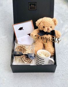 a brown teddy bear sitting in a black box next to a cup and some gold rings