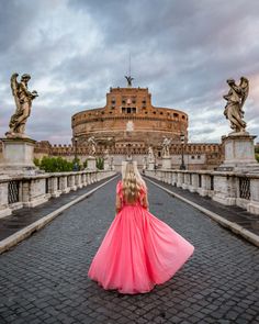 a woman in a pink dress is standing on a bridge with statues and buildings behind her