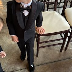 a little boy in a tuxedo standing next to a chair