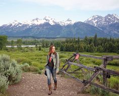 a woman walking down a dirt road next to a wooden fence with mountains in the background