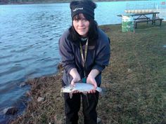 a woman is holding a fish in her hands by the water's edge while wearing a beanie