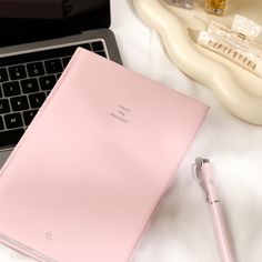 a pink notebook sitting on top of a desk next to a laptop computer and pen