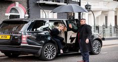 a man standing next to a woman holding an umbrella in front of a black range rover