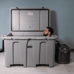 a man with a fake head sitting in a large gray cooler next to a trash can