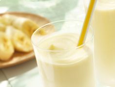 two glasses filled with orange juice next to sliced bananas on a plate and another glass has a straw in it