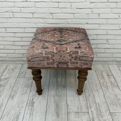 an old wooden stool with a patterned upholstered seat sits against a white brick wall