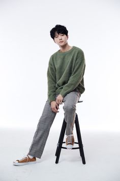 a young man sitting on top of a stool wearing a green sweater and grey pants