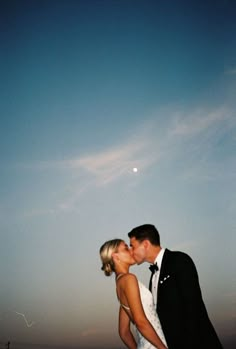 a bride and groom kissing in front of the moon on their wedding day at sunset