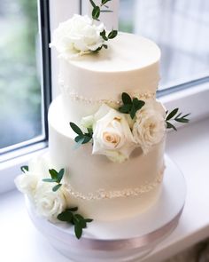 a three tiered white wedding cake with flowers on the top and bottom, sitting in front of a window