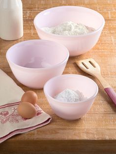 three pink bowls with eggs and flour in them on a wooden table next to utensils