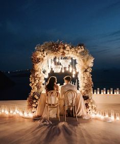 two people sitting at a table in front of candles and an arch decorated with flowers