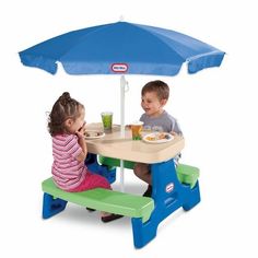 two children sitting at a little tikes table with an umbrella over them and one child eating