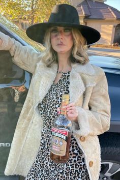 a woman in a leopard print dress and black hat holding a bottle of liquor while standing next to a car