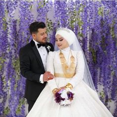 a man and woman dressed in wedding attire standing next to each other with purple flowers behind them