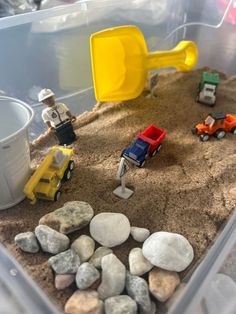 a plastic container filled with rocks and toy cars on top of sand next to a bucket