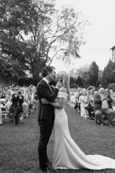 a bride and groom kissing in front of an audience at their outdoor wedding ceremony, black and white photograph