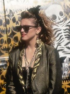 a woman wearing sunglasses and a jacket standing in front of a wall with skulls on it