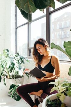 a woman sitting on a window sill reading a book in front of potted plants