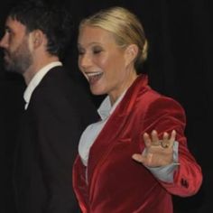 a woman in a red velvet jacket and white shirt is smiling at the camera while another man stands behind her