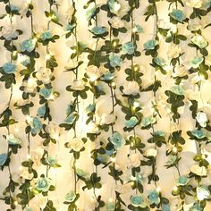 white flowers and green leaves are hanging from a curtain with lights in the corner behind them