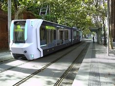 an electric train traveling down the tracks next to a tree lined sidewalk with people walking on it