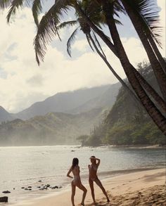 two women in bathing suits walking on the beach next to palm trees and mountain range