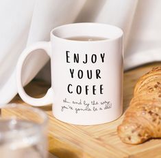 a white coffee mug sitting on top of a wooden table next to a croissant