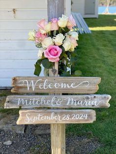 a wooden welcome sign with flowers in the center on grass next to a white house