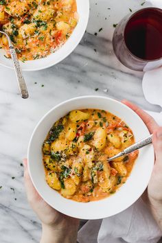 two hands holding bowls of pasta and garnished with parsley next to a glass of red wine