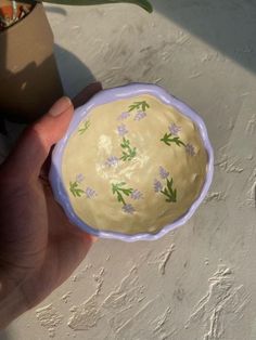 a hand holding a yellow and green bowl with flowers on it, next to a potted plant