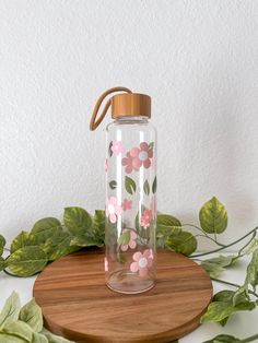 a glass water bottle with pink flowers on it sitting on a wooden table next to green leaves