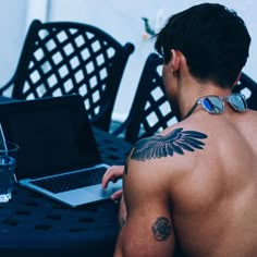 a man with tattoos on his back sitting at a table using a laptop and drinking water