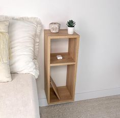 a small wooden shelf next to a bed with pillows and a plant on the side