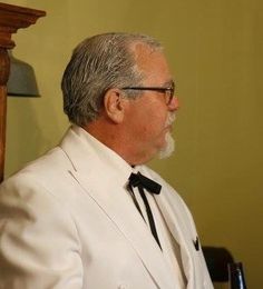 an older man in a white suit and black tie sitting at a table with a glass of wine