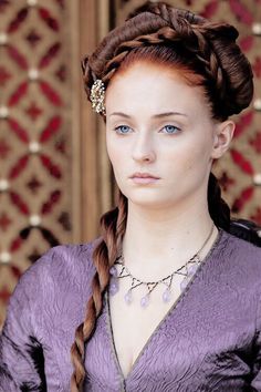 a woman with long braids and blue eyes wearing a purple dress in front of a wall