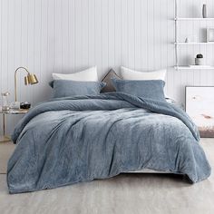 a bed with blue comforter and pillows in a white room next to a book shelf