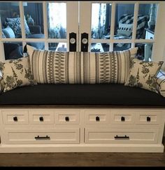 a white bench with drawers and pillows in front of two large windows on the wall