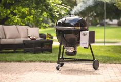 a black grill sitting on top of a brick patio next to a green lawn and trees