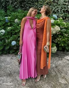 two women standing next to each other in front of some bushes and flowers with their heads together