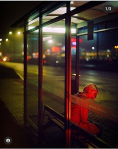 a person sitting on a bench in front of a bus stop at night with their head turned to the side