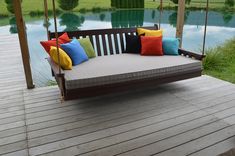 a wooden swing with pillows on it sitting next to a body of water in the background