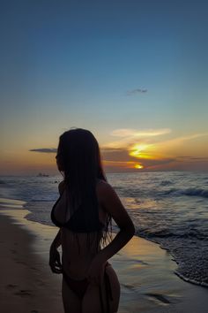 a woman standing on top of a beach next to the ocean at sun set with her back turned