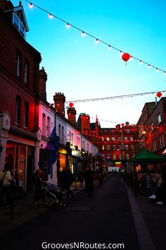 people are walking down the street in an old town at night with lights strung above them